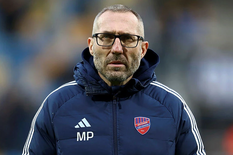 Marek Papszun Mężczyzna z krótkimi włosami, brodą i okularami ubrany w granatową sportową kurtkę z inicjałami MP oraz herbem klubu sportowego, stoi na tle rozmytego stadionu.