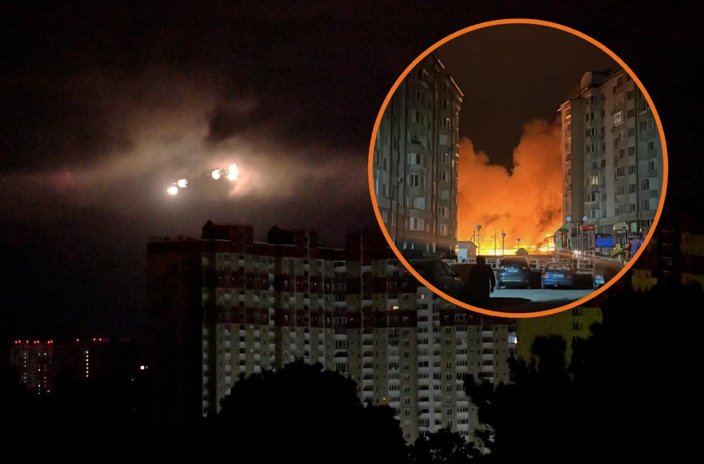 Nocna panorama miasta z wysokimi blokami, za którymi unosi się intensywna łuna ognia i kłęby dymu. W okrągłym powiększeniu widoczny wyraźnie pożar między budynkami, ogień oświetla całą ulicę i budynki mieszkalne.