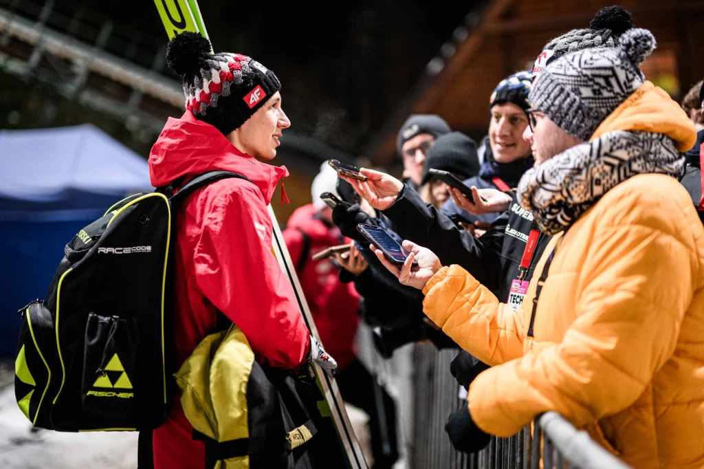 Sportowiec w czerwonej kurtce i czapce udziela wywiadu dziennikarzom po zawodach narciarskich, trzyma narty i plecak. Wokół niego grupka osób z mikrofonami oraz aparatami, wszyscy ubrani w zimową odzież.