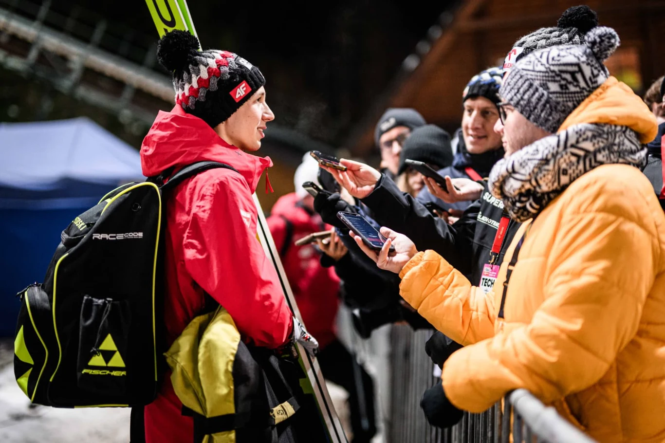 Kacper Tomasiak Sportowiec w czerwonej kurtce i czapce udziela wywiadu dziennikarzom po zawodach narciarskich, trzyma narty i plecak. Wokół niego grupka osób z mikrofonami oraz aparatami, wszyscy ubrani w zimową odzież.