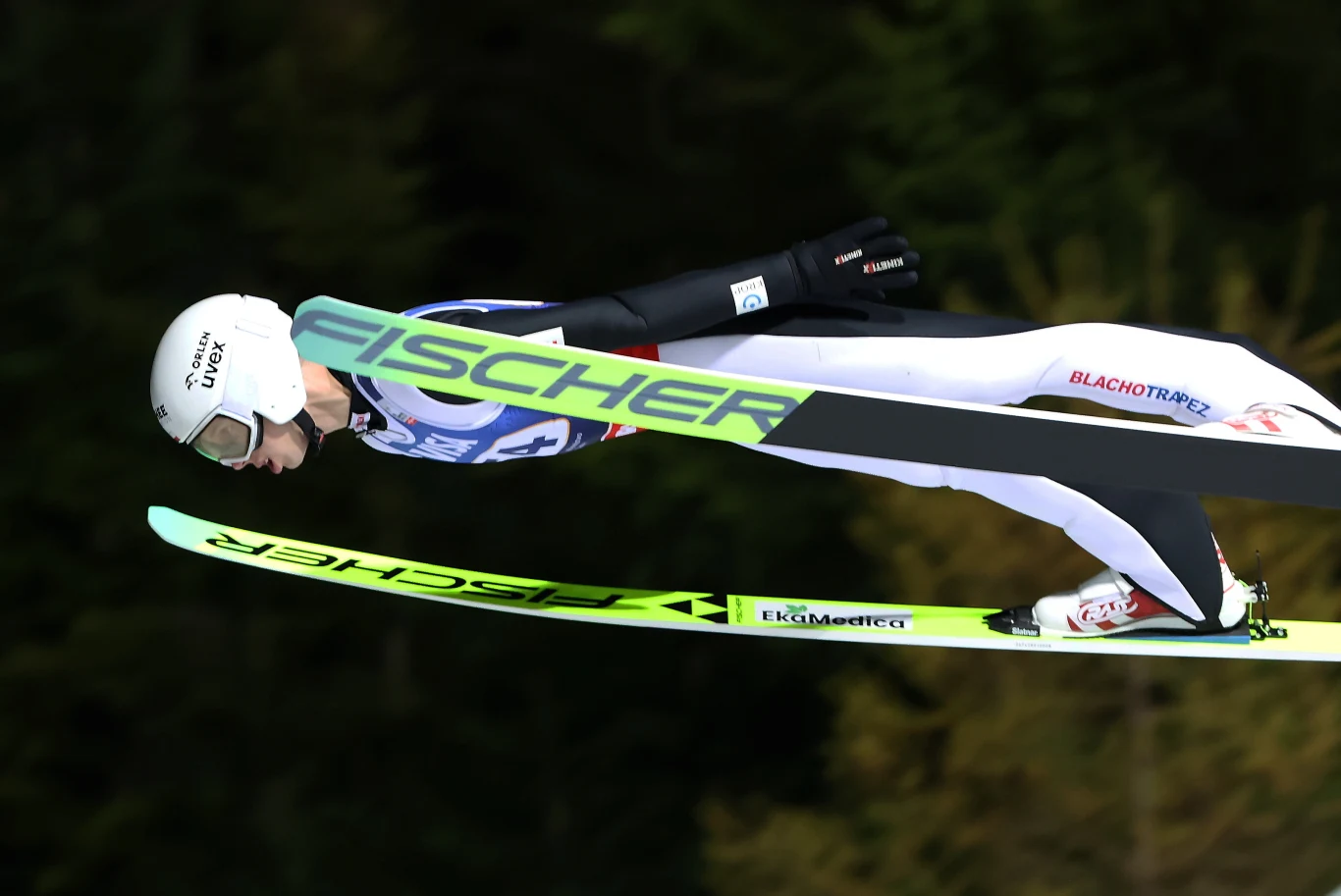 Zawodnik w kombinezonie narciarskim wykonuje lot skokiem narciarskim, lecąc nad stokiem na nartach, w tle rozmyte drzewa.