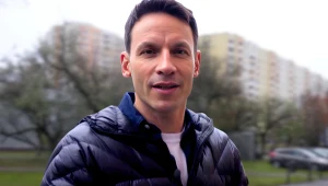 Młody mężczyzna w ciemnej kurtce, w tle osiedle bloków mieszkaniowych