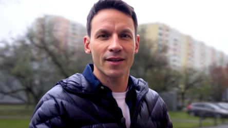 Młody mężczyzna w ciemnej kurtce, w tle osiedle bloków mieszkaniowych