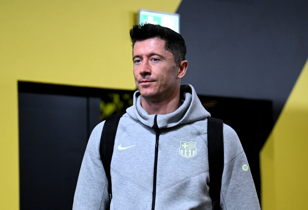 Mężczyzna ubrany w szarą bluzę z herbem FC Barcelona oraz czarny plecak stoi na tle kontrastujących żółto-czarnych ścian.