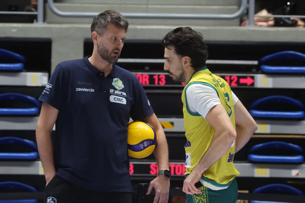 Michał Winiarski i Miguel Tavares, rozgrywający Aluron CMC Warty Zawiercie Trener oraz zawodnik podczas rozmowy na hali sportowej, obok piłka do siatkówki, w tle puste ławki rezerwowych.