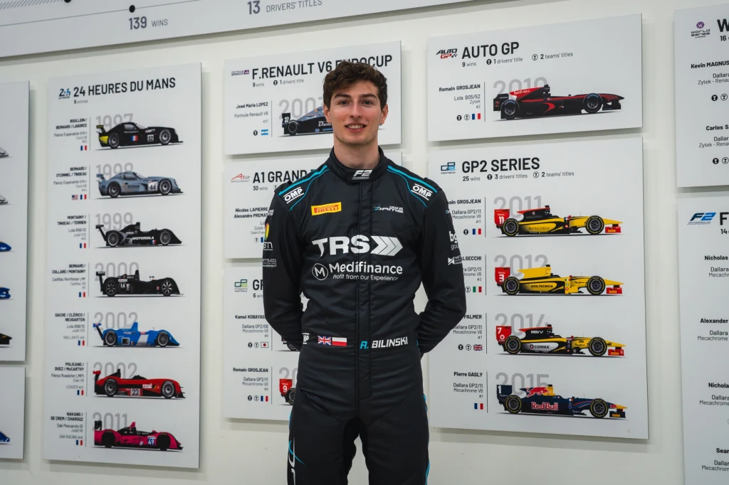 Roman Biliński awansował do Formuły 2 Młody mężczyzna w czarnym kombinezonie wyścigowym z logo sponsorów stoi na tle plansz z ilustracjami bolidów i informacjami o różnych seriach wyścigowych.