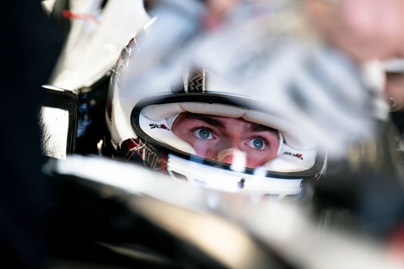 Zbliżenie na twarz kierowcy wyścigowego w kasku siedzącego w kokpicie bolidu, skupione spojrzenie skierowane przed siebie, otaczają go elementy techniczne pojazdu.
