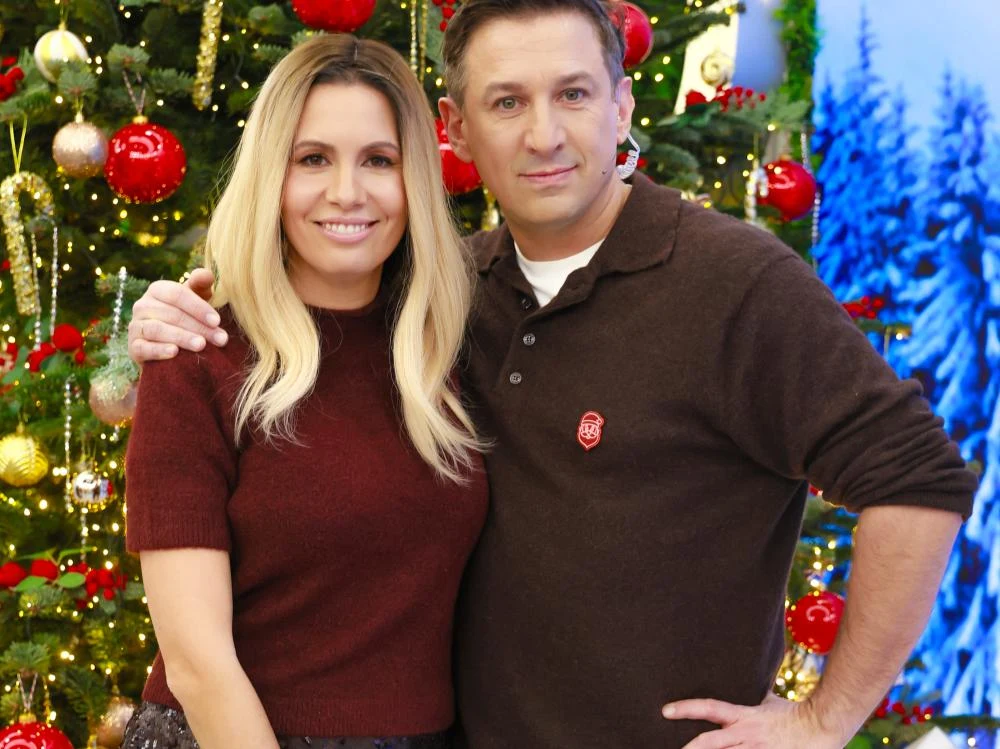 Agnieszka Hyży, Maciej Rock Para uśmiechniętych osób ubranych w swetry stoi razem przed świątecznie udekorowaną choinką z czerwonymi i złotymi ozdobami. W tle widać dekoracje bożonarodzeniowe i elementy nawiązujące do zimowego klimatu.