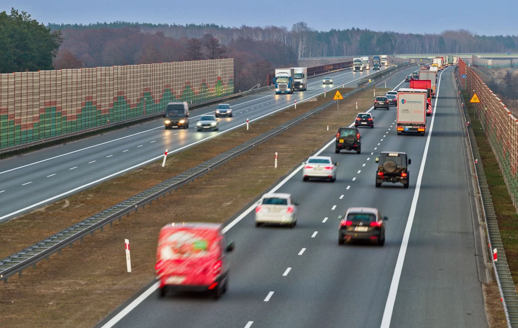 Ruchliwa autostrada z dużą liczbą samochodów osobowych i ciężarowych, po obu stronach drogi ekrany dźwiękochłonne, widoczne oznaczenia drogowe i fragmenty lasu w tle.