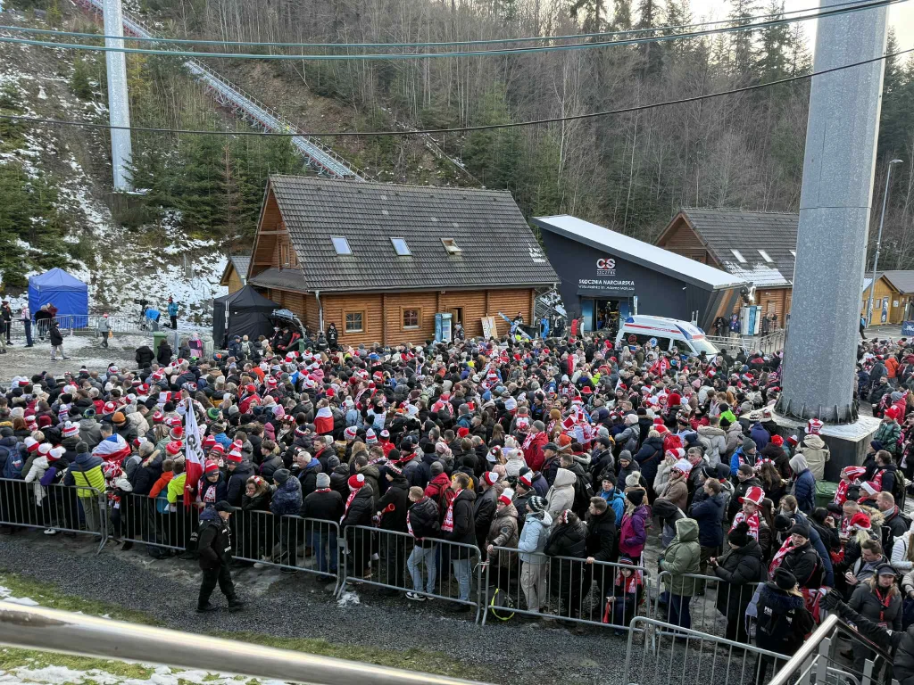 Tłum ludzi ubranych w zimowe ubrania zgromadzony za metalowymi barierkami przed wejściem do dużego budynku z drewnianym dachem, w tle las i nowoczesna infrastruktura sportowa, wiele osób ma na głowach czerwone czapki.