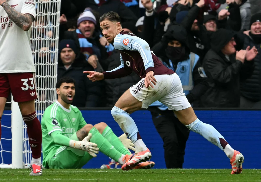 Zawodnik drużyny piłkarskiej w bordowo-niebieskim stroju świętuje zdobycie gola, biegnąc przed bramką, podczas gdy bramkarz drużyny przeciwnej w zielonym komplecie siedzi na ziemi ze zdziwioną miną. Kibice w tle żywo reagują na sytuację na boisku.