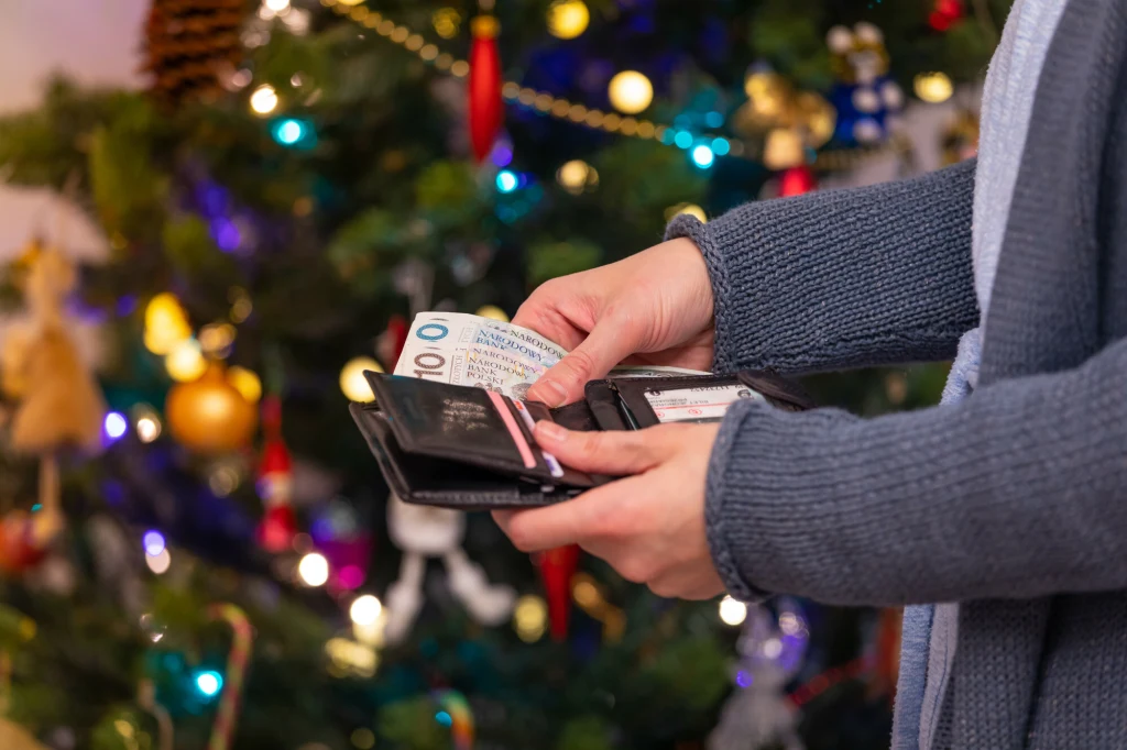 Polacy wydadzą więcej pieniędzy na nadchodzące Święta niż w ubiegłym roku Ręce trzymające portfel z widocznymi banknotami i kartami, tło stanowi ozdobiona świątecznymi dekoracjami choinka.