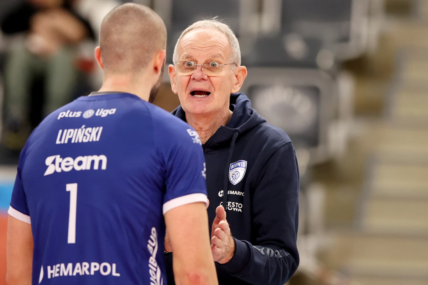 Trener wyrażający silne emocje podczas rozmowy z zawodnikiem siatkarskim w niebieskim stroju z nazwiskiem Lipiński na plecach, w tle widoczne rozmyte trybuny.