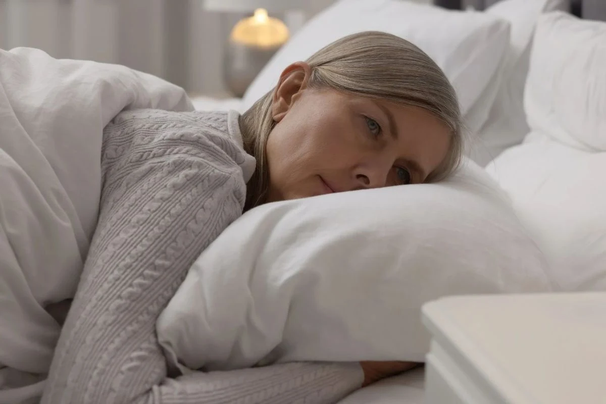 Kobieta w średnim wieku leżąca w łóżku, oparta głową na poduszce, z zamyślonym wyrazem twarzy, ubrana w jasny sweter, w tle widoczna lampka nocna.