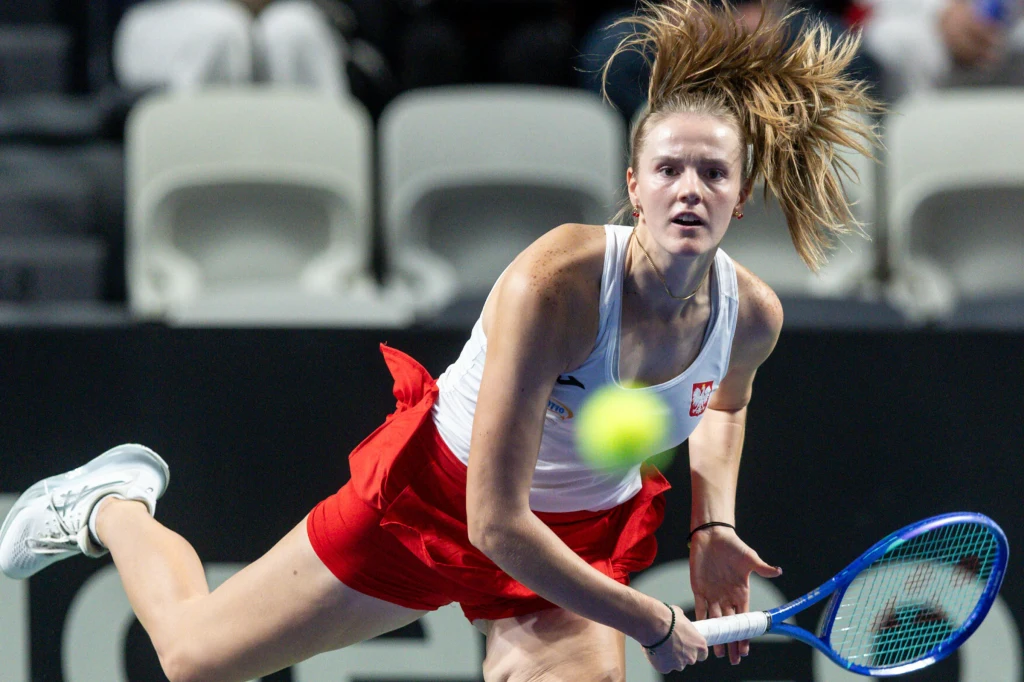 Tenisistka w białym topie i czerwonej spódniczce wykonuje dynamiczny ruch rakietą podczas meczu, widoczne skupienie na twarzy, piłka tenisowa w ruchu, puste trybuny w tle.