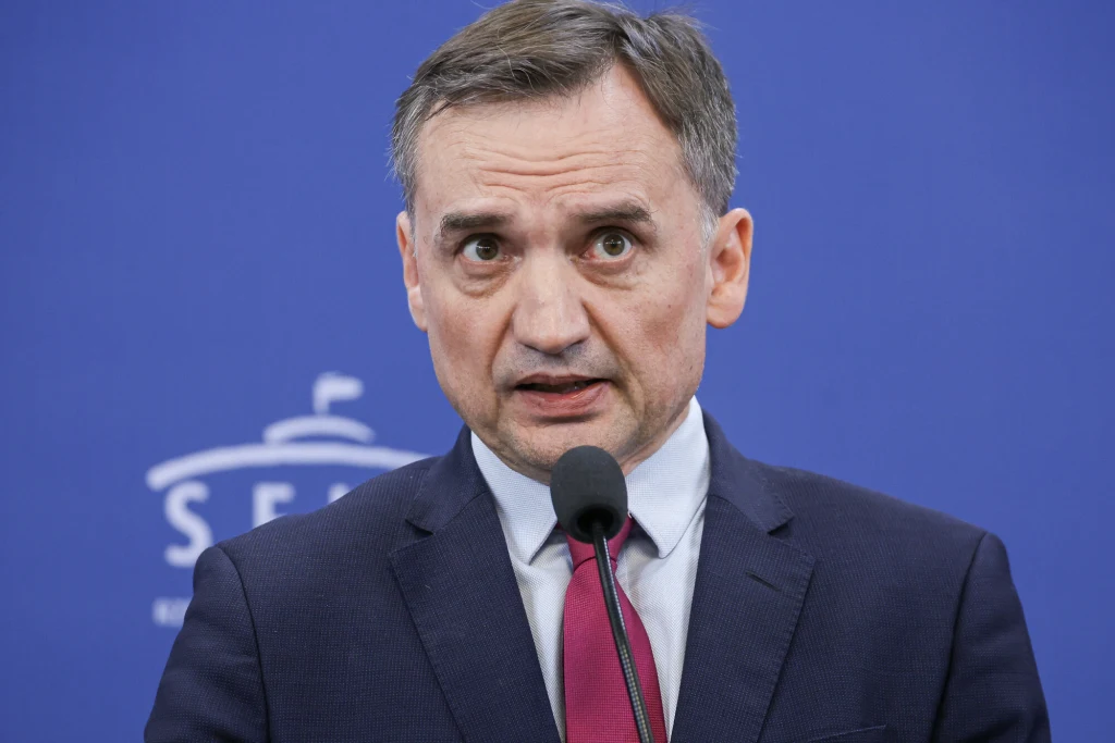 Zbigniew Ziobro Mężczyzna w garniturze i czerwonym krawacie stoi przed mikrofonem na tle niebieskiej ścianki z napisem oraz symbolem kopuły, patrzy poważnie w stronę kamery.