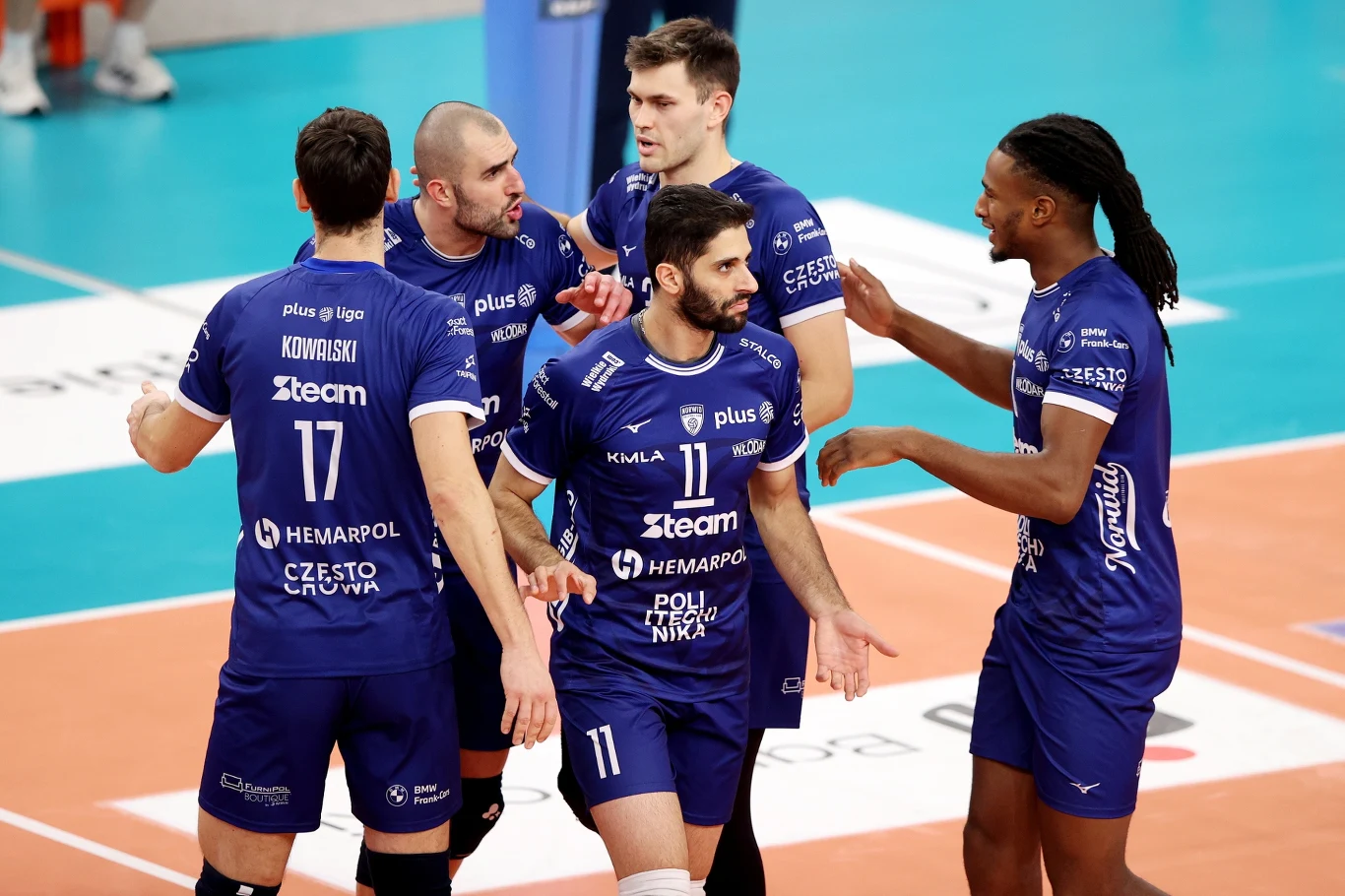 Grupa siatkarzy w niebieskich strojach drużynowych świętuje wspólnie zdobycie punktu na boisku do siatkówki, zbierając się wokół siebie i okazując emocje oraz sportową jedność.