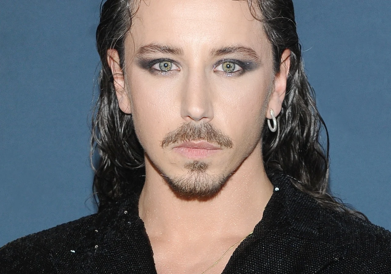 Michał Szpak Mężczyzna z zarostem, długimi, ciemnymi włosami i wyrazistym makijażem wokół oczu, ubrany w czarną marynarkę z delikatnie połyskującego materiału, na tle jednolitej, niebieskiej ściany.