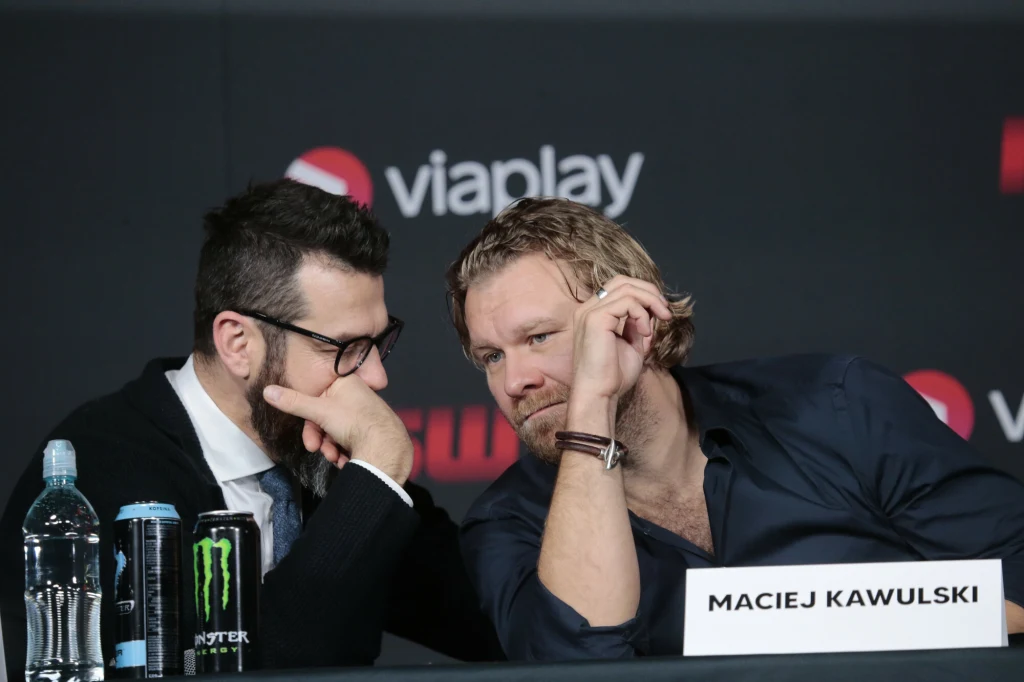 Martin Lewandowski i Maciej Kawulski, szefowie KSW Dwóch mężczyzn siedzi obok siebie przy stole konferencyjnym i rozmawia półszeptem, jeden z nich podpiera głowę dłonią, przed nimi tabliczka z napisem 'Maciej Kawulski', na stole butelka wody, napój energetyczny i mikrofon, w tle logo Viaplay.