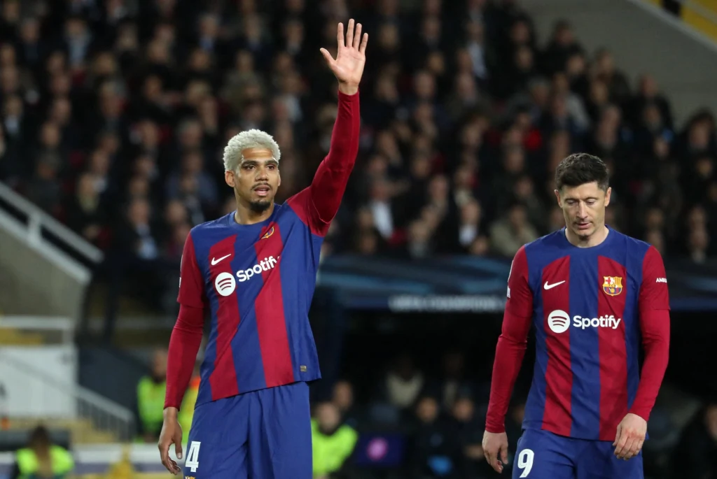 Dwóch piłkarzy drużyny FC Barcelona w granatowo-bordowych strojach na tle stadionu, jeden z zawodników unosi rękę, drugi stoi obok z opuszczoną głową.