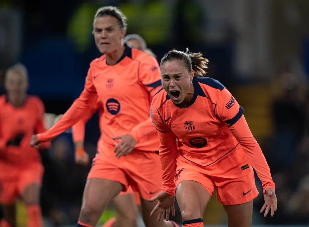 Ewa Pajor celebrująca bramkę Dwie piłkarki w pomarańczowych strojach drużyny rozgrywają mecz, jedna z nich wyraża silne emocje, prawdopodobnie po ważnym momencie gry, stadion oświetlony w tle.