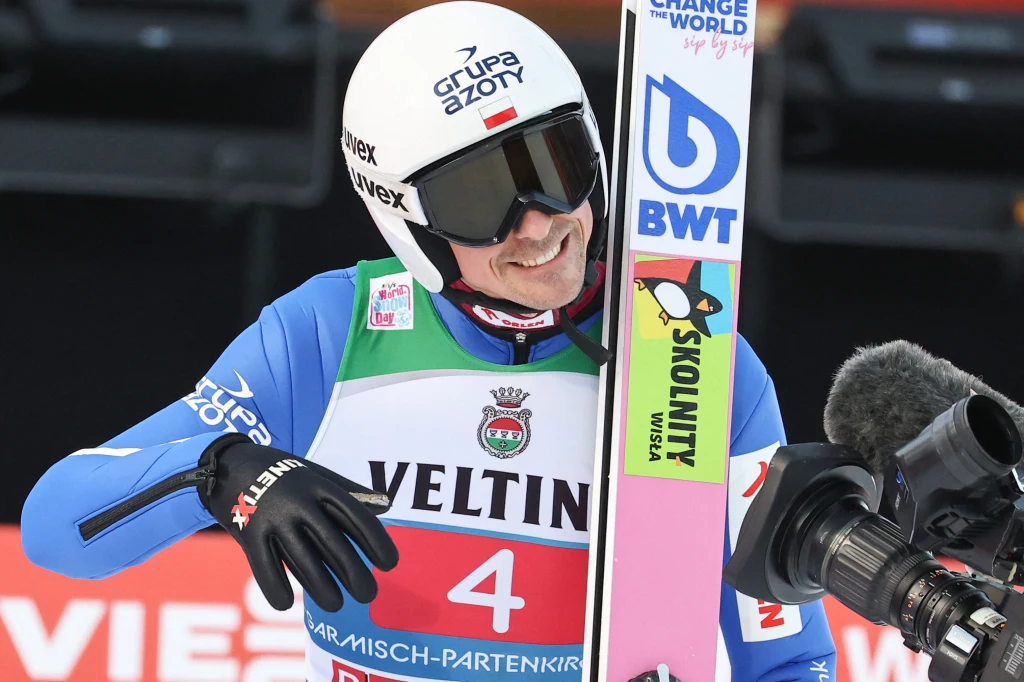 Sportowiec ubrany w strój narciarski z charakterystycznym kaskiem i goglami trzyma narty, uśmiecha się i spogląda w stronę kamery, w tle widoczne bannery z logotypami sponsorów.