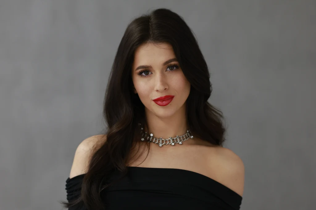 Roxie Kobieta o długich ciemnych włosach, elegancko ubrana w czarną, odkrywającą ramiona suknię, z subtelnym makijażem i czerwonymi ustami, nosi srebrny naszyjnik na tle szarego, neutralnego tła.