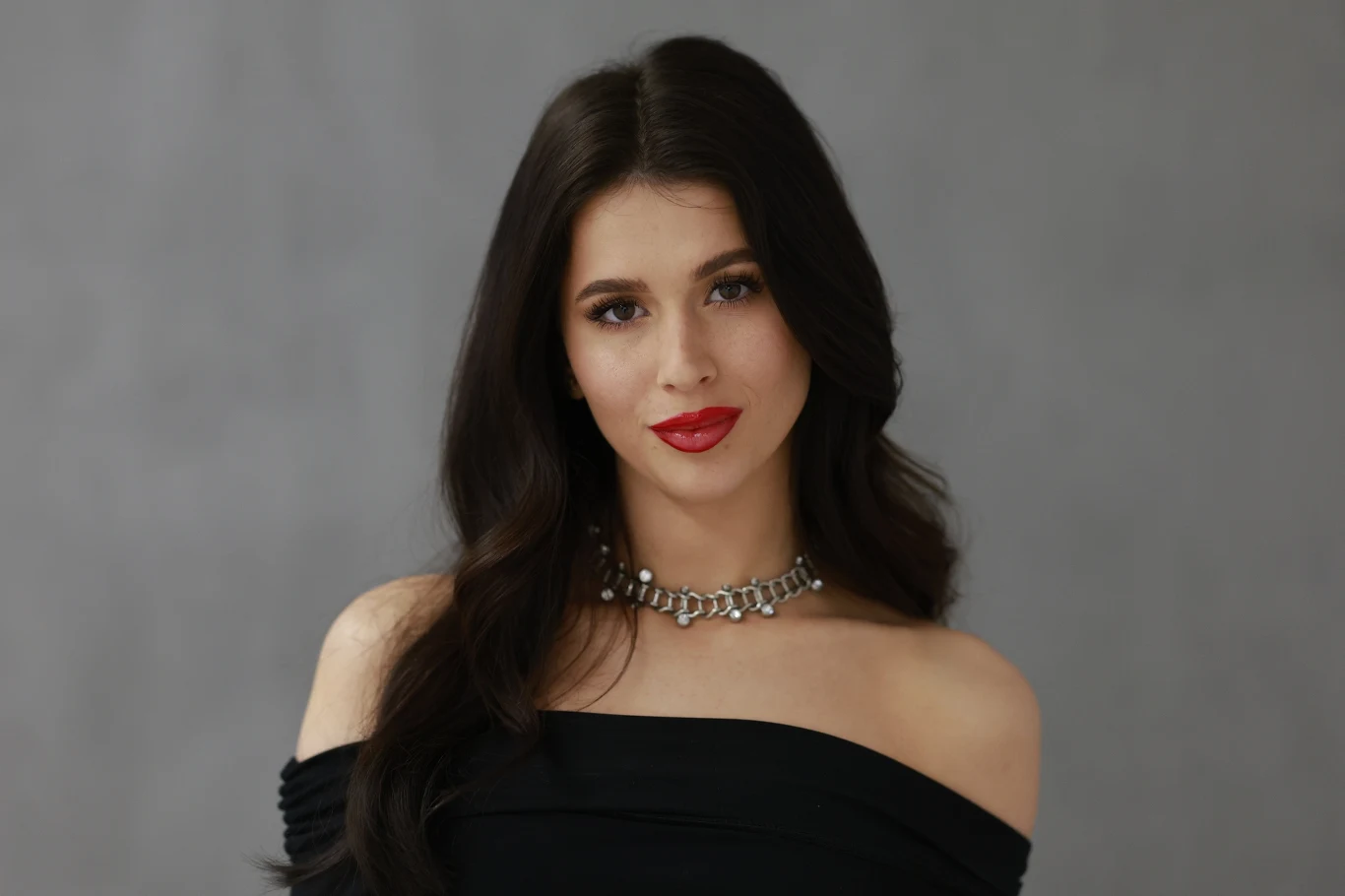 Roxie Kobieta o długich ciemnych włosach, elegancko ubrana w czarną, odkrywającą ramiona suknię, z subtelnym makijażem i czerwonymi ustami, nosi srebrny naszyjnik na tle szarego, neutralnego tła.