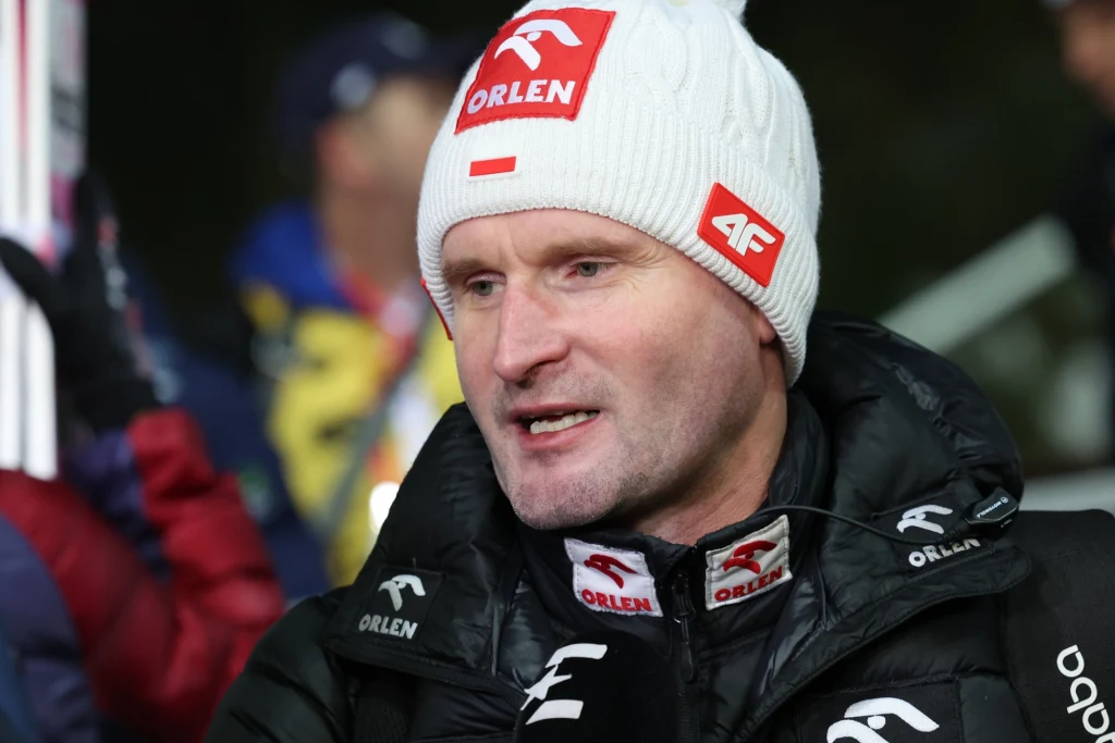 Maciej Maciusiak Mężczyzna w białej czapce z logo Orlen oraz 4F, ubrany w czarną kurtkę, udzielający wywiadu. W tle zamazana postać i elementy sprzętu sportowego, możliwie podczas wydarzenia sportowego zimą.