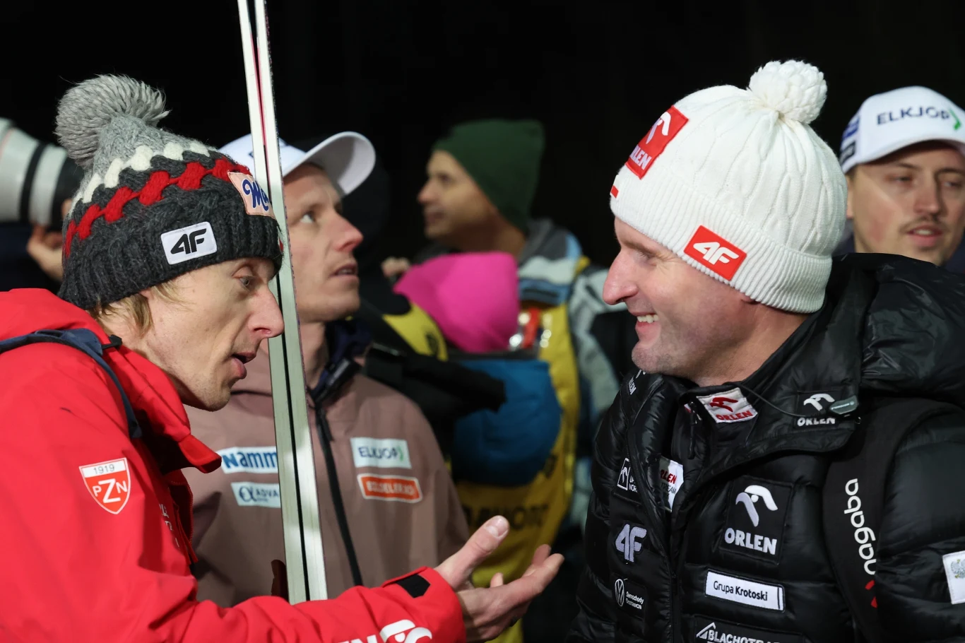 Dwóch mężczyzn ubranych w sportowe zimowe kurtki i czapki rozmawia ze sobą, obok nich stoją inni uczestnicy wydarzenia, a w tle tłum ludzi i ciemne niebo.