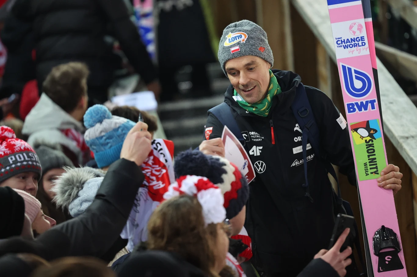 Skoczek narciarski w stroju sportowym i z nartą rozdaje autografy oraz wita się z grupą kibiców ubranych w biało-czerwone czapki i szaliki.