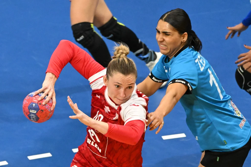 Intensywna walka podczas meczu piłki ręcznej kobiet, zawodniczka w czerwono-białym stroju próbuje przedostać się z piłką obok rywalki w niebieskiej koszulce, obie wykazują duże zaangażowanie i determinację.