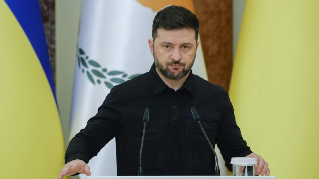 Prezydent Ukrainy Wołodymyr Zełenski Mężczyzna w ciemnej koszuli stoi za mównicą, przed mikrofonami, w tle widoczne są flagi Ukrainy oraz innego państwa.