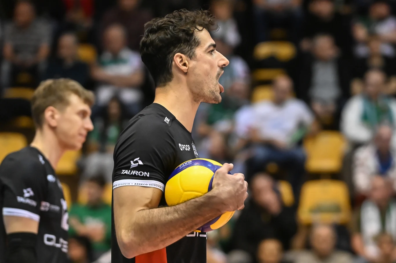 Siatkarz w czarnej koszulce trzymający piłkę do siatkówki pod pachą, w tle widoczny rozmazany tłum kibiców na trybunach.