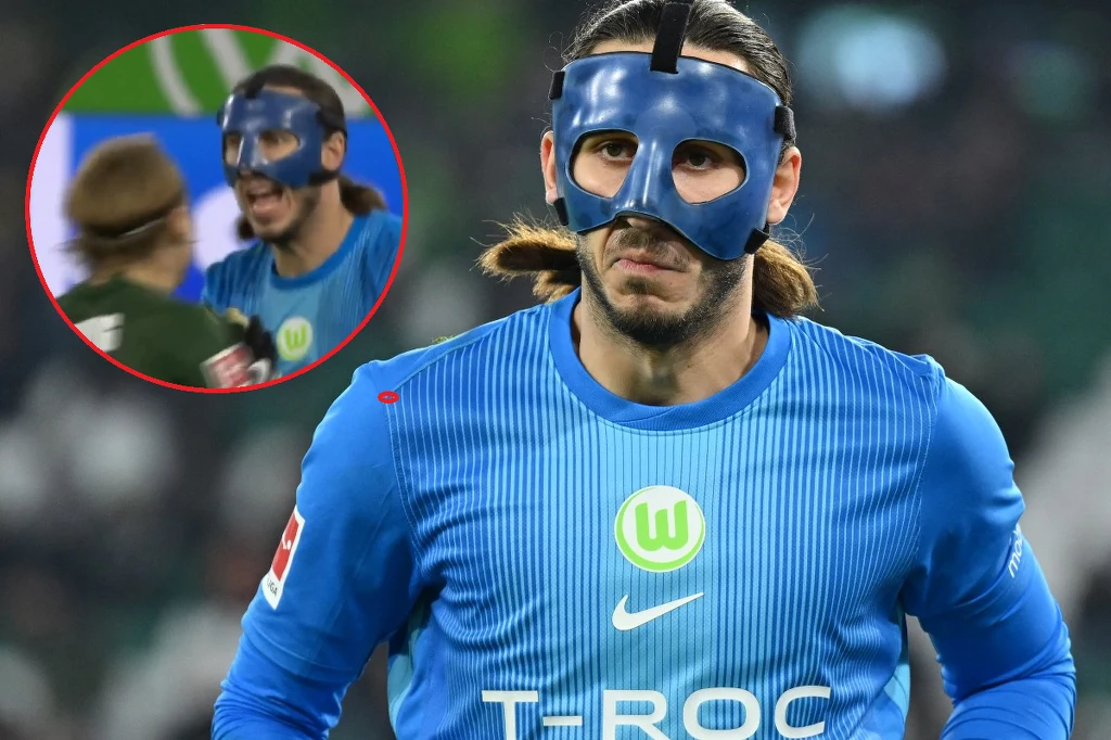 Kamil Grabara Piłkarz w niebieskiej koszulce z logiem drużyny VfL Wolfsburg i napisem sponsora T-ROC, mający na twarzy ochraniaczą maskę sportową; w lewym górnym rogu widoczne powiększenie, na którym widać emocjonalną wymianę zdań z innym zawodnikiem.