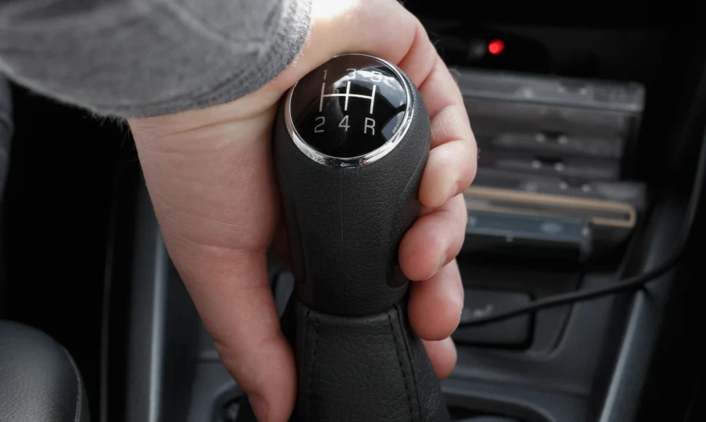 Hamowanie silnikiem to zwykle zalecana metoda wytracania prędkości Ręka trzymająca gałkę manualnej skrzyni biegów w samochodzie z widocznym schematem przełożeń oraz elementami konsoli środkowej.