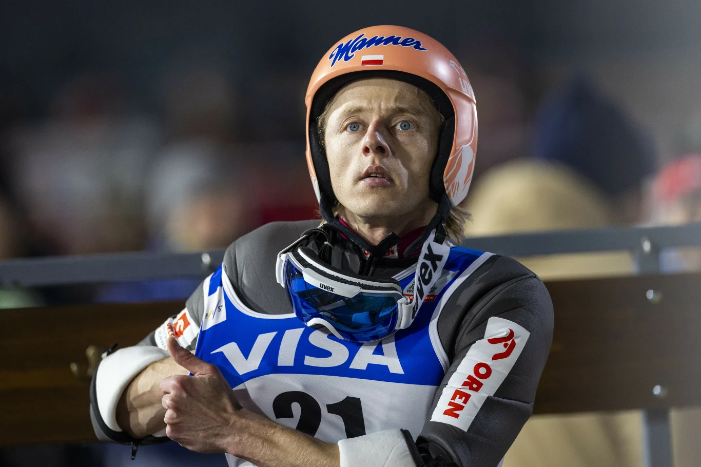Dawid Kubacki Skoczek narciarski w kombinezonie startowym, kasku i goglach trzyma ręce skrzyżowane na klatce piersiowej, z widocznym napięciem oczekuje na swoją kolej. W tle rozmyci kibice i elementy skoczni.
