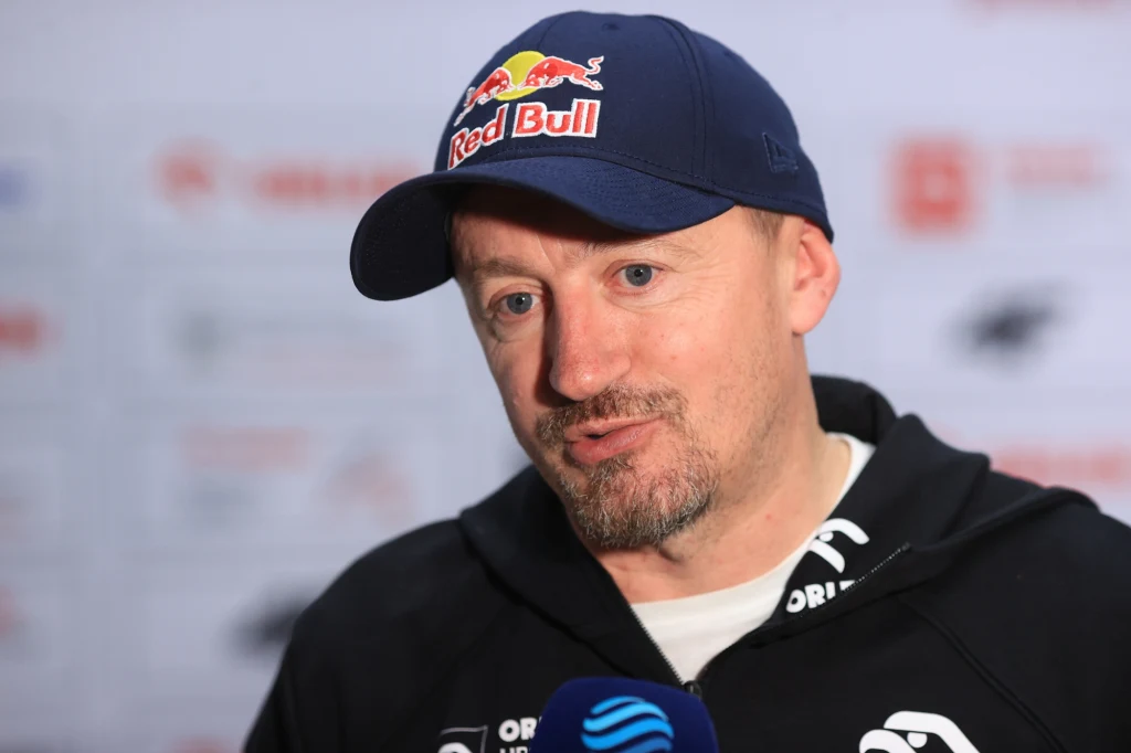 Adam Małysz Mężczyzna w granatowej czapce Red Bull, ubrany w sportową bluzę, udziela wywiadu, trzymając przy ustach mikrofon. W tle rozmyte logotypy oraz białe tło z napisami.