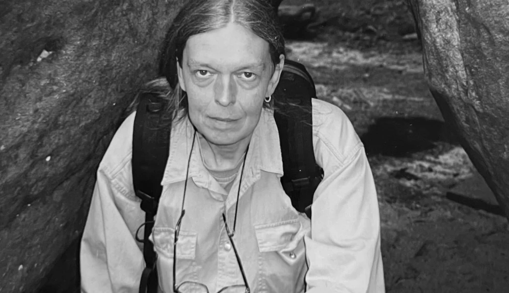 Kobieta z długimi, siwymi włosami i plecakiem na ramionach wśród dużych skał, ubrana w jasną koszulę, z poważnym wyrazem twarzy.