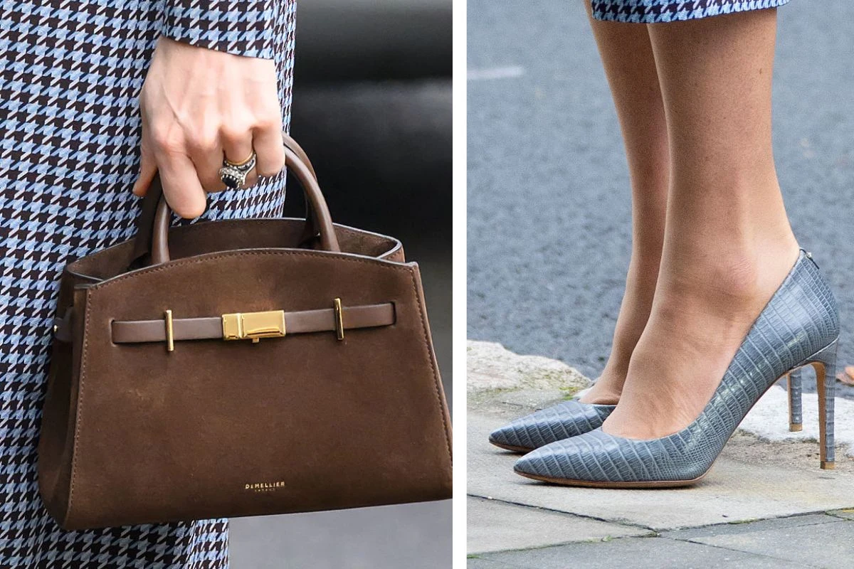 Brązowa torebka trzymana w lewej ręce ubranej w ubranie w pepitkę, obok szare szpilki na wysokim obcasie na tle chodnika, również przy stroju w pepitkę.