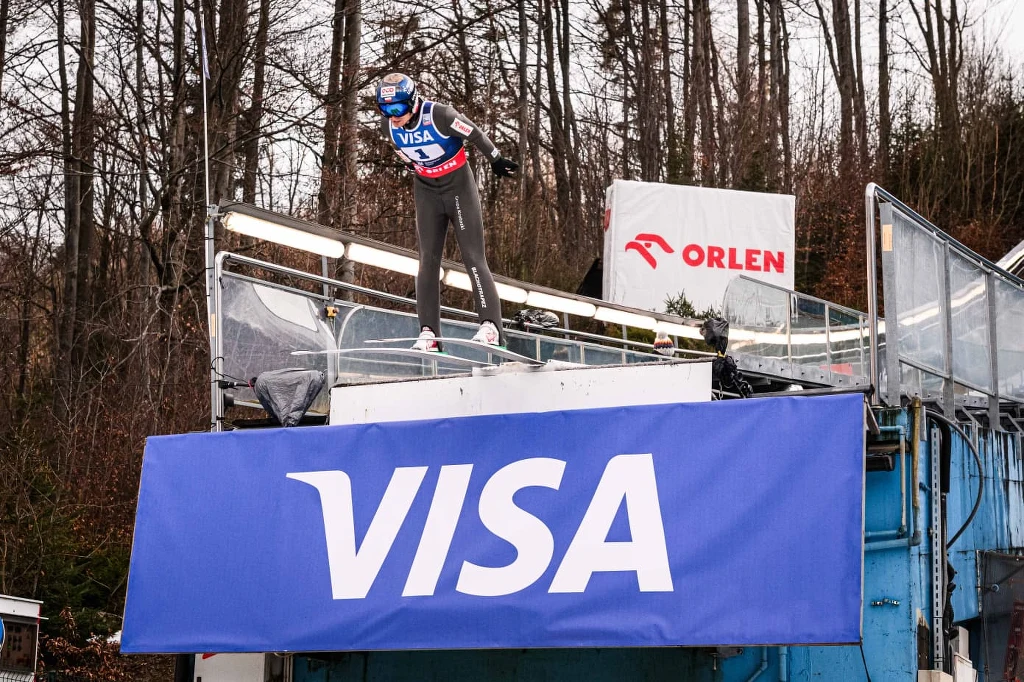 Maciej Kot Zawodnik w kombinezonie narciarskim przygotowujący się do skoku narciarskiego stoi na progu skoczni, w tle widoczne są drzewa oraz reklamy sponsorskie VISA i ORLEN.