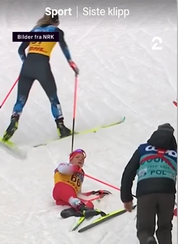 Zawodniczka w stroju narciarskim leży na śniegu, najwyraźniej po upadku podczas biegu narciarskiego, obok niej dwie inne osoby: jedna ubrana na sportowo stoi na nartach, druga w kurtce z napisem POL nadchodzi z pomocą.