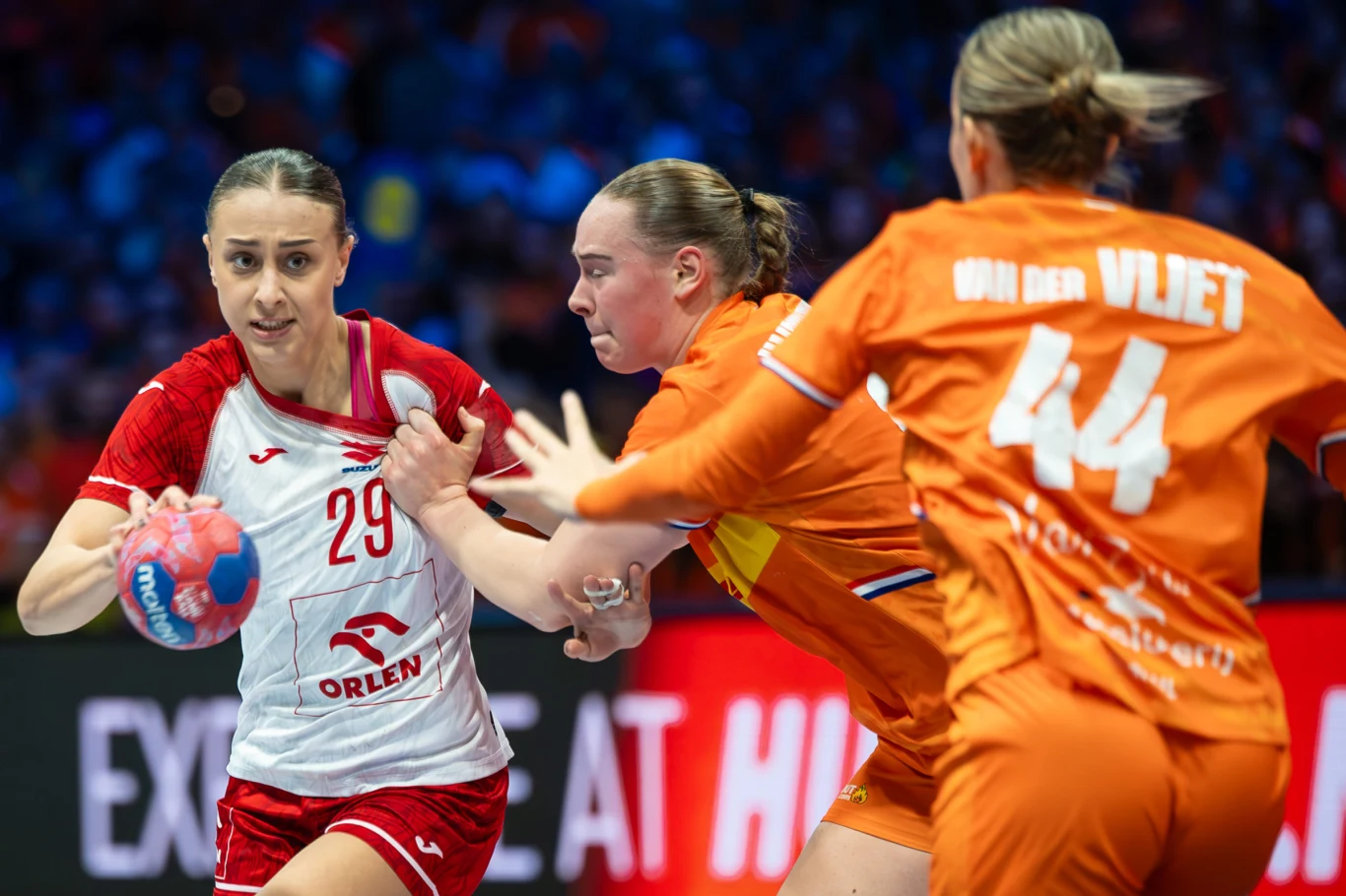 Katarzyna Cygan Zawodniczka w biało-czerwonym stroju z piłką próbuje przejść przez defensywę dwóch przeciwniczek ubranych w pomarańczowe stroje podczas intensywnego momentu meczu piłki ręcznej.