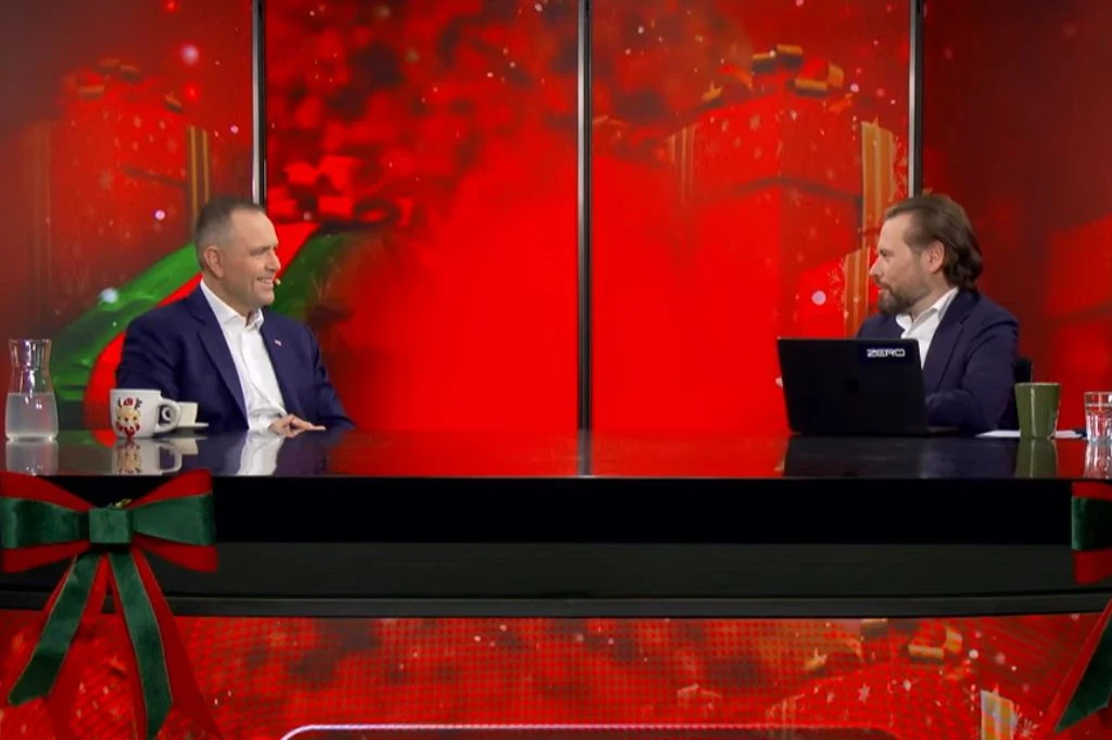 Dwóch mężczyzn siedzi naprzeciw siebie przy szklanym stole w profesjonalnym studiu telewizyjnym z czerwonym, świątecznym tłem. Na stole znajdują się filiżanki, dzbanek z wodą i laptop. Dekoracja z zieloną kokardą sugeruje tematykę świąteczną.
