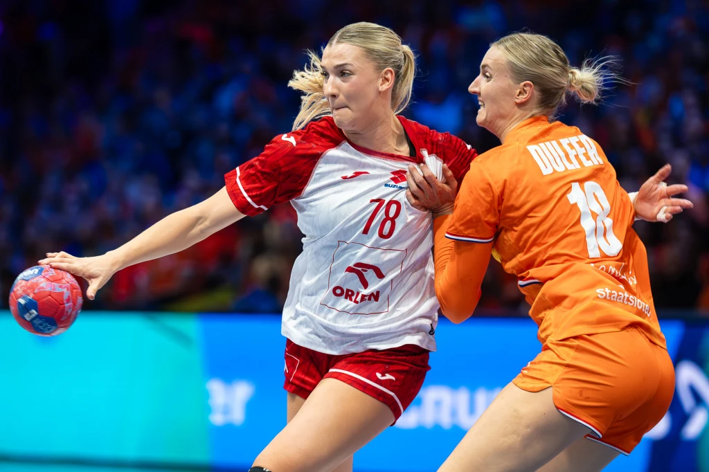 Dwie zawodniczki grające w piłkę ręczną, jedna w czerwono-białym stroju z piłką w ręku próbuje przebić się przez obronę przeciwniczki ubranej w pomarańczowy strój. Intensywny moment sportowej walki na boisku.