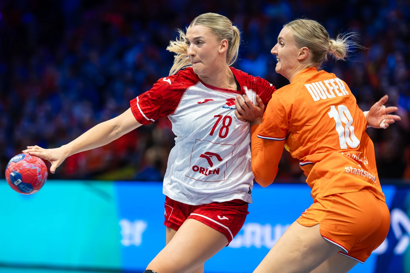 Paulina Uścinowicz Dwie zawodniczki grające w piłkę ręczną, jedna w czerwono-białym stroju z piłką w ręku próbuje przebić się przez obronę przeciwniczki ubranej w pomarańczowy strój. Intensywny moment sportowej walki na boisku.