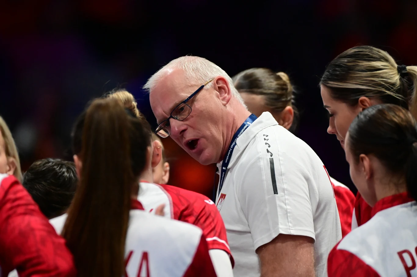 Arne Senstad Trener drużyny narodowej w białej koszulce pochylony w kierunku zawodniczek w czerwono-białych strojach, energicznie udziela wskazówek w trakcie przerwy podczas meczu siatkówki.