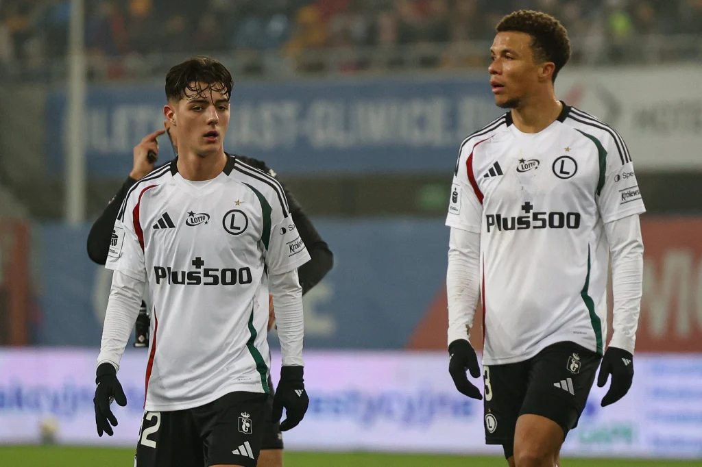 Piast Gliwice - Legia Warszawa Dwóch piłkarzy drużyny Legia Warszawa w białych strojach po meczu, widoczna publiczność na trybunach w tle, na strojach logo sponsora oraz klubowe emblematy.