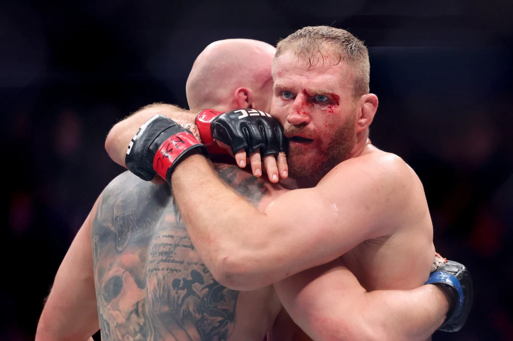 Jan Błachowicz zremisował z Bogdanem Guskovem Dwóch zawodników MMA w trakcie walki, przytulających się podczas starcia. Jeden z nich ma rozcięty łuk brwiowy i ślady krwi na twarzy, obaj noszą rękawice ochronne. Atmosfera rywalizacji sportowej i napięcia.