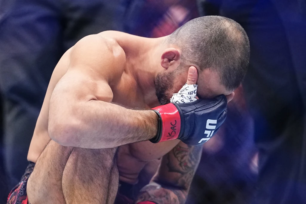 Alexandre Pantoja na UFC 323 Zawodnik MMA w rękawicach z logo UFC przykuca w klatce, zakrywając twarz dłonią w geście rozczarowania lub zmęczenia. Wokół widoczne rozmyte postacie sędziów lub obsługi walki.